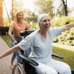 Joyful woman with cancer wearing a headscarf, smiling with arms outstretched in a wheelchair, being pushed by a caregiver outdoors. The uplifting moment highlights the healing journey and emotional support often found in cannabis-assisted cancer care.