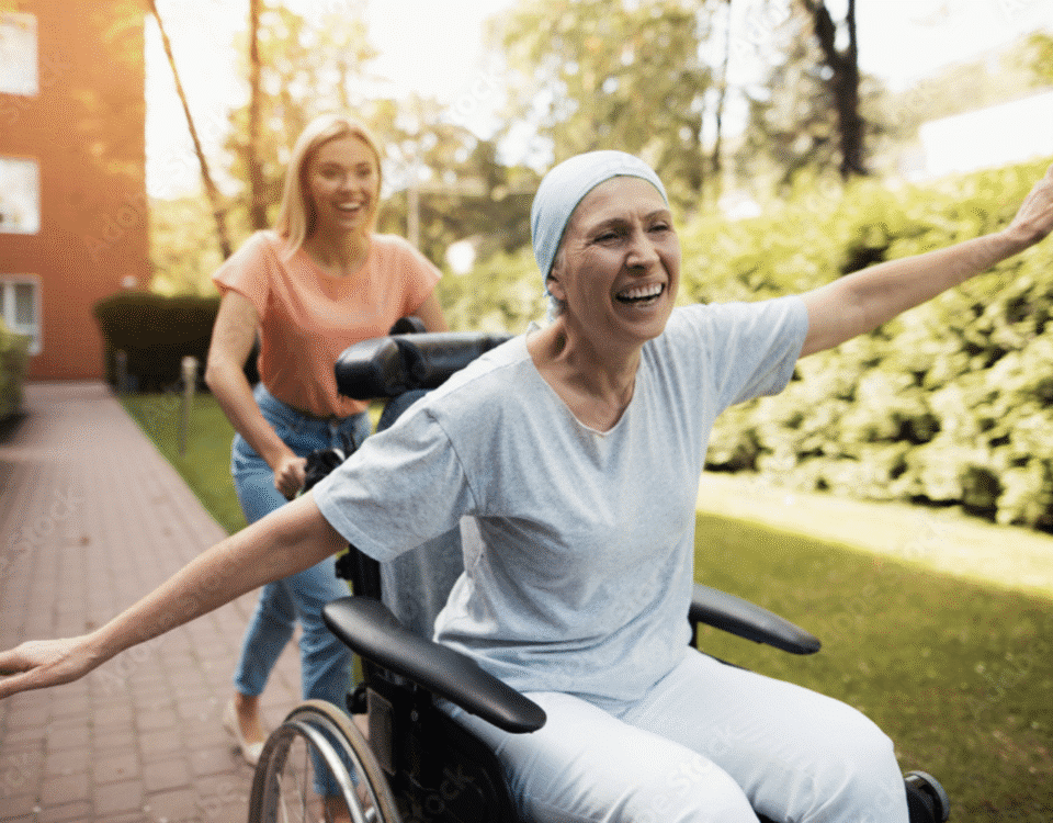 Joyful woman with cancer wearing a headscarf, smiling with arms outstretched in a wheelchair, being pushed by a caregiver outdoors. The uplifting moment highlights the healing journey and emotional support often found in cannabis-assisted cancer care.