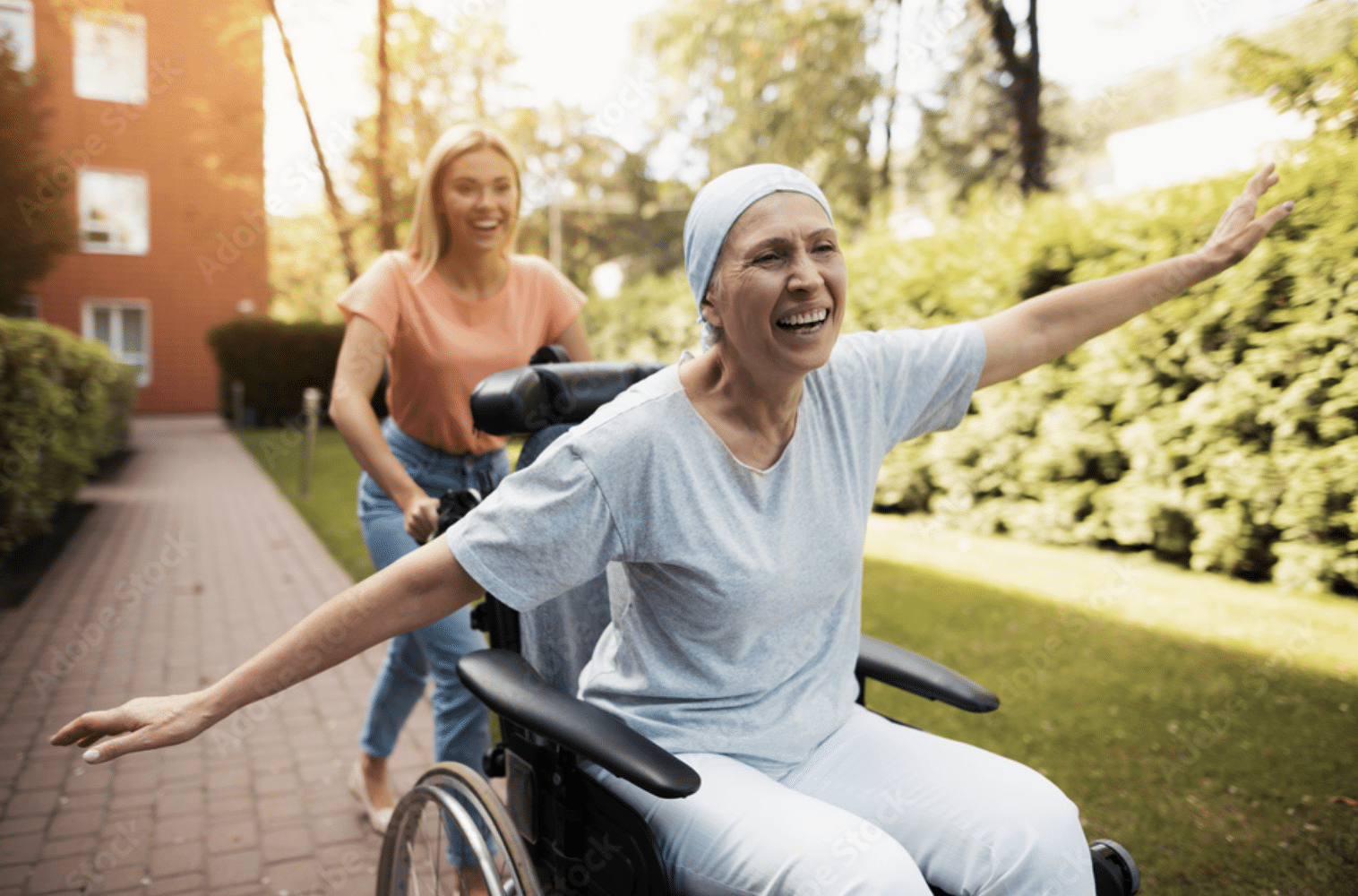 Joyful woman with cancer wearing a headscarf, smiling with arms outstretched in a wheelchair, being pushed by a caregiver outdoors. The uplifting moment highlights the healing journey and emotional support often found in cannabis-assisted cancer care.